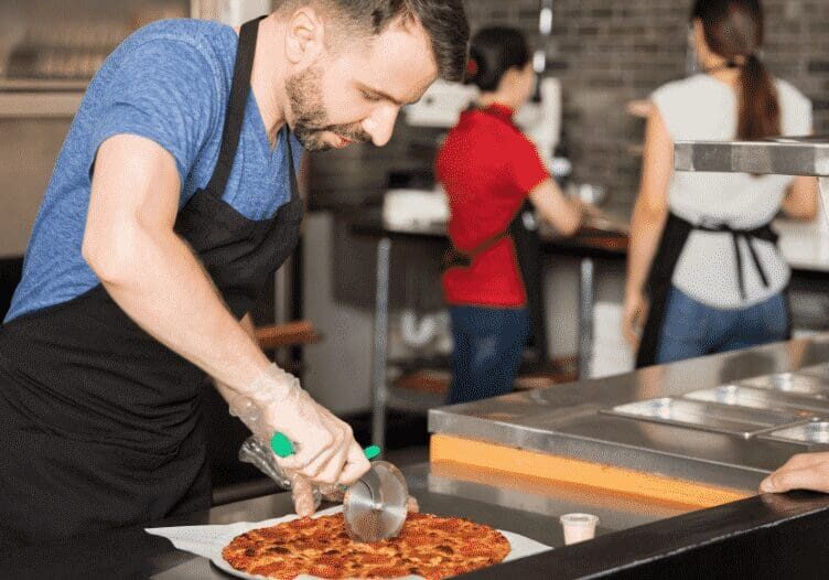 Man slicing pizza in a restaurant kitchen.